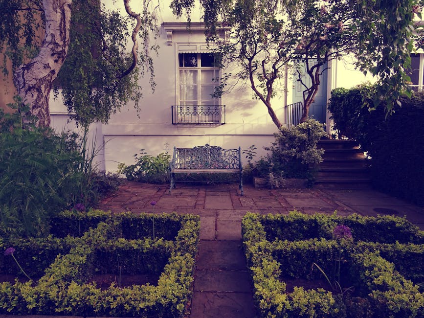A quaint cobblestone courtyard lined with white townhouses on either side, featuring potted plants, small trees, and shrubs along the building facades. Some of the buildings have black iron balconies,